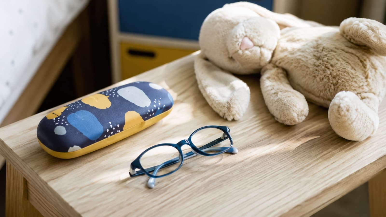 Petites lunettes rondes d’enfant posées sur une table en bois clair à côté de crayons de couleur et d’un doudou, lumière douce