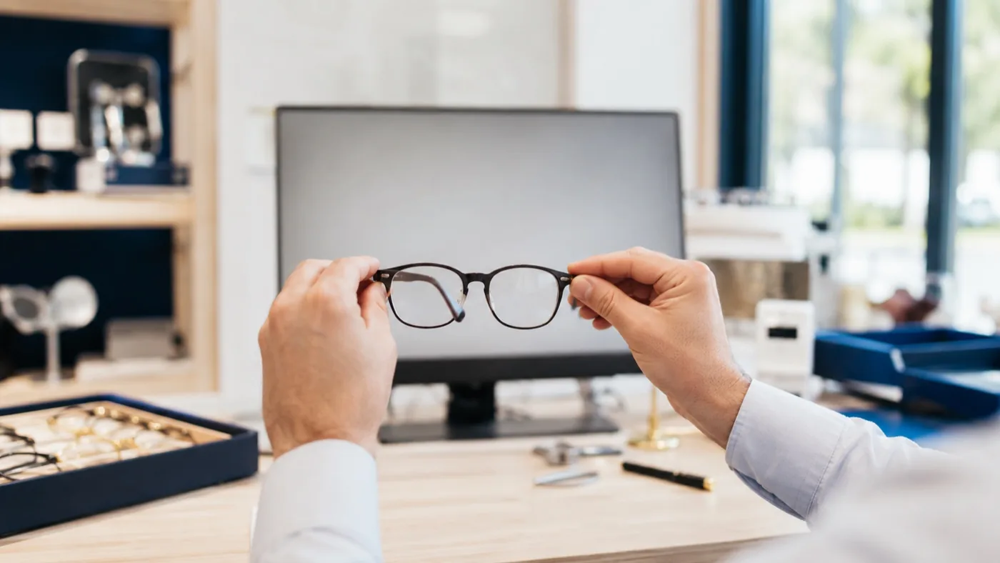 Un opticien tient une paire de lunettes devant un écran d’ordinateur pour vérifier la vision de proximité en boutique